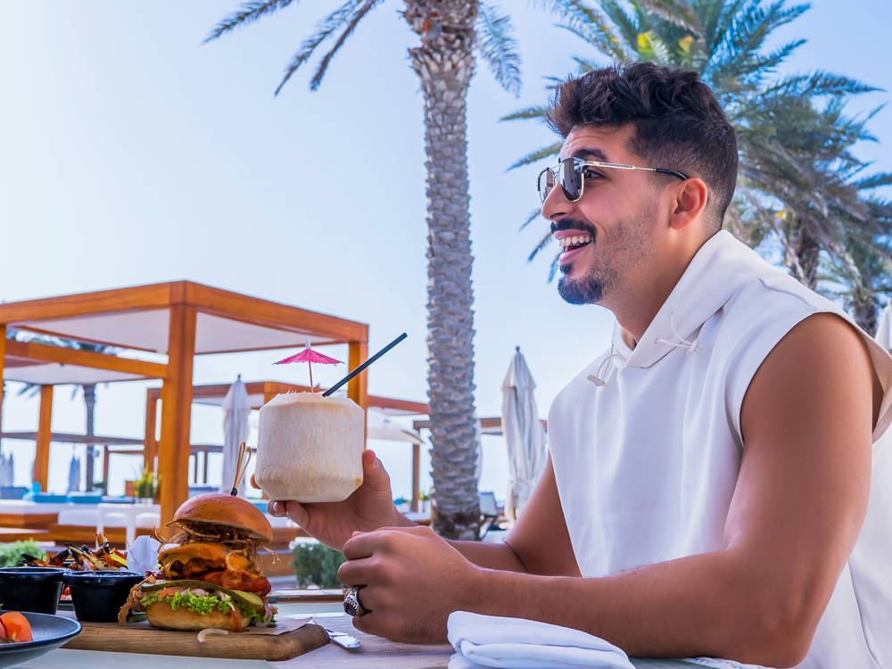 Man by a cabana at Saadiyat Beach Club enjoying a burger and a refreshing coconut cocktail in a relaxed setting.