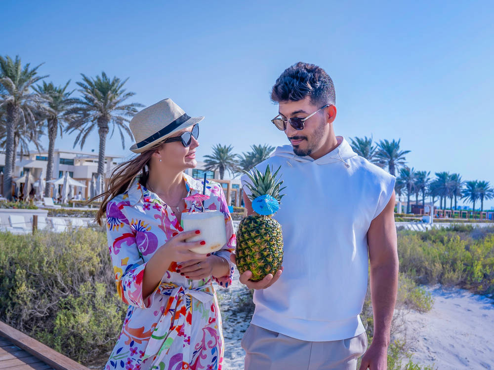 At Saadiyat Beach Club, a couple enjoys cocktails in a coconut and pineapple with cabanas and palm trees in the background.