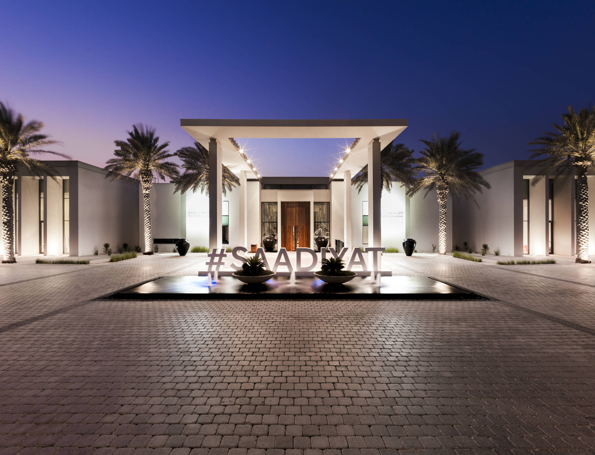 Saadiyat Beach Club at dusk. The modern and white palm tree lined building has "#SAADIYAT" in front of it